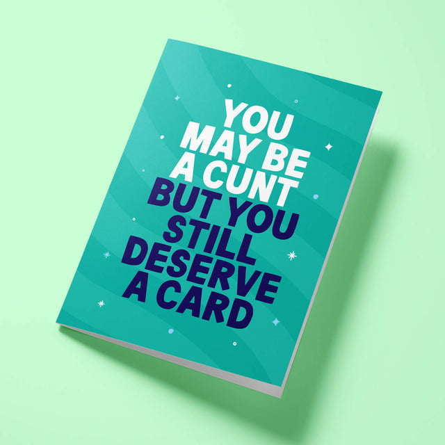 Green 'You Still Deserve A Card' rude birthday card laid flat at an angle on a bright background, with dark navy 'BUT YOU STILL DESERVE A CARD' lettering standing out against the white text.