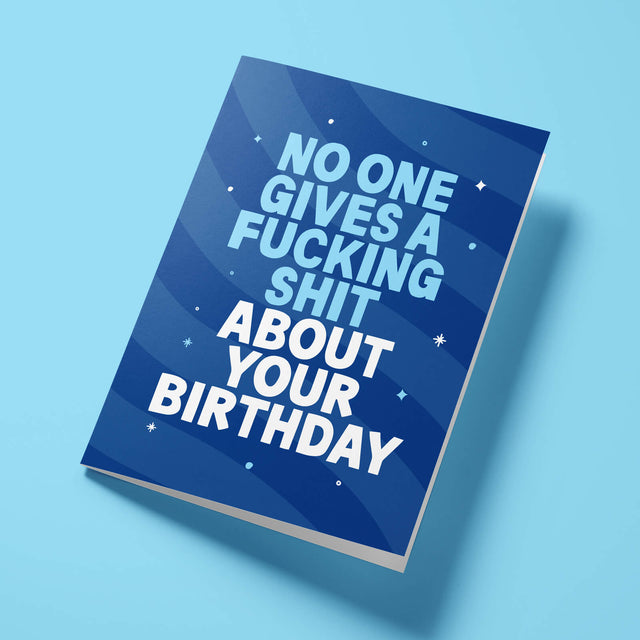 Navy blue 'No One Gives A Shit' rude birthday card laid flat at an angle on a bright background, with light blue 'NO ONE GIVES A FUCKING SHIT' lettering standing out against the white text.