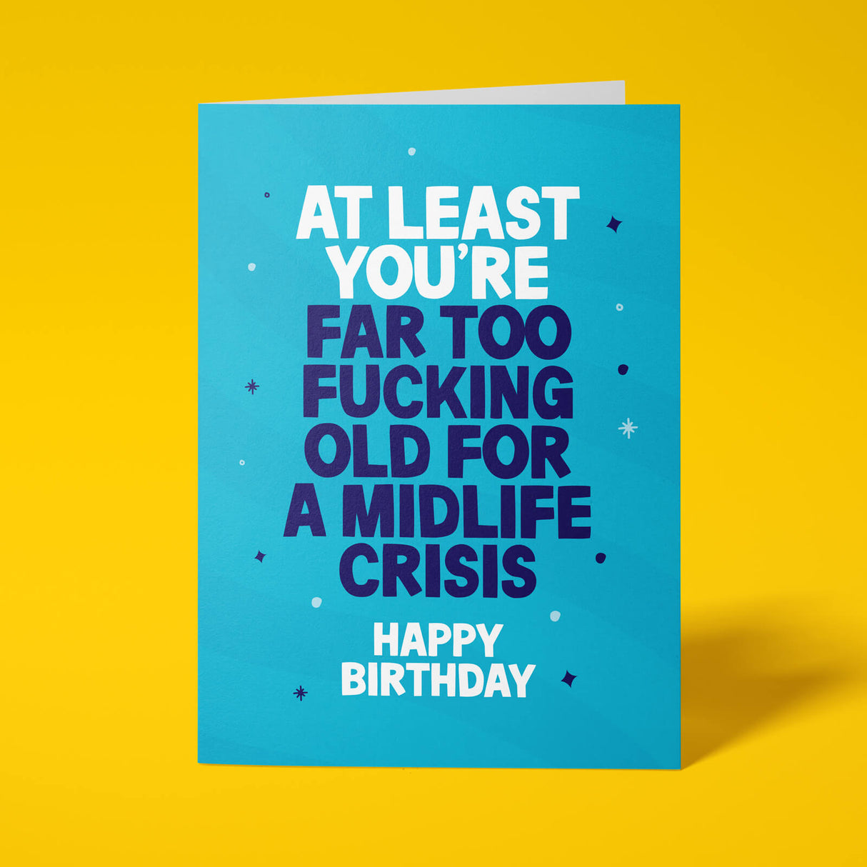 Bright teal A5 card displayed against a styled coloured background. Bold white and dark navy chunky typography reads 'AT LEAST YOU'RE FAR TOO FUCKING OLD FOR A MIDLIFE CRISIS HAPPY BIRTHDAY'. Starburst ray pattern in teal with scattered sparkle and dot details.