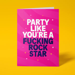 Hot pink rude birthday card standing upright on a bright background. Bold white and dark navy chunky typography reads 'PARTY LIKE YOU'RE A FUCKING ROCK STAR' over pink wave stripes with yellow and white sparkle details.