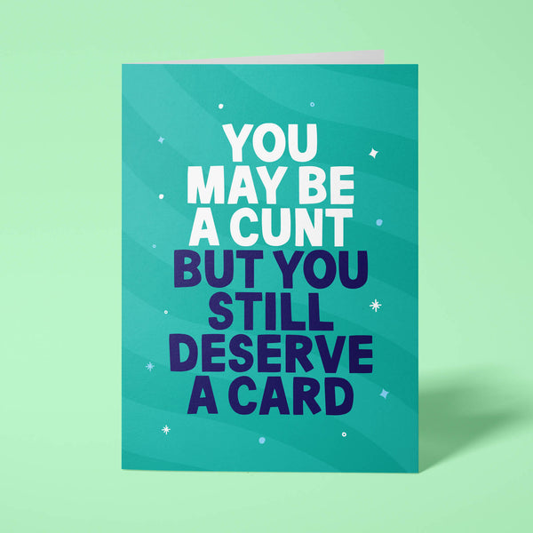 Green rude birthday card standing upright on a bright background. Bold white and dark navy chunky typography reads 'YOU MAY BE A CUNT BUT YOU STILL DESERVE A CARD' over Green wave stripes with white sparkle details.