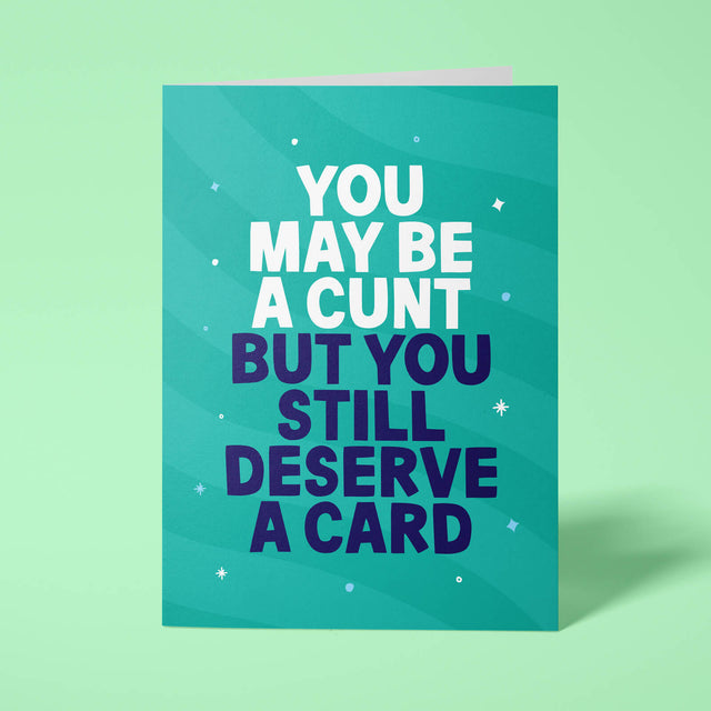 Green rude birthday card standing upright on a bright background. Bold white and dark navy chunky typography reads 'YOU MAY BE A CUNT BUT YOU STILL DESERVE A CARD' over Green wave stripes with white sparkle details.
