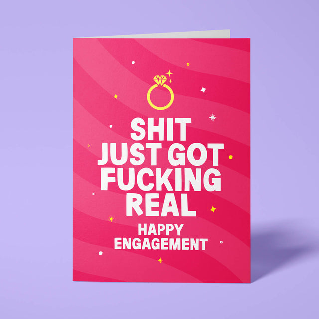 Raspberry pink rude engagement card standing upright on a bright background. Bold white chunky typography reads 'SHIT JUST GOT FUCKING REAL HAPPY ENGAGEMENT' beneath pink wave stripes with a gold diamond ring illustration and yellow and white sparkle details.
