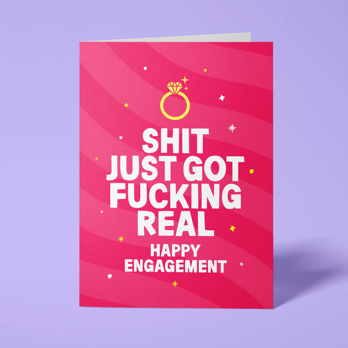 Raspberry pink rude engagement card standing upright on a bright background. Bold white chunky typography reads 'SHIT JUST GOT FUCKING REAL HAPPY ENGAGEMENT' beneath pink wave stripes with a gold diamond ring illustration and yellow and white sparkle details.