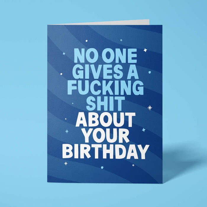 Navy blue rude birthday card standing upright on a bright background. Bold light blue and white chunky typography reads 'NO ONE GIVES A FUCKING SHIT ABOUT YOUR BIRTHDAY' over navy wave stripes with white and light blue sparkle details.
