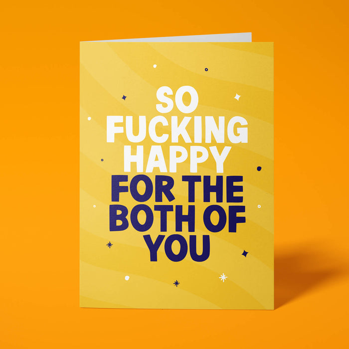 Golden yellow rude congratulations card standing upright on a bright background. Bold white and dark navy chunky typography reads 'SO FUCKING HAPPY FOR THE BOTH OF YOU' over yellow wave stripes with white and navy sparkle details.