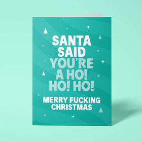 Green rude Christmas card standing upright on a bright background. Bold white and pale mint chunky typography reads 'SANTA SAID YOU'RE A HO! HO! HO! MERRY FUCKING CHRISTMAS' over green wave stripes with white Christmas tree and sparkle details.