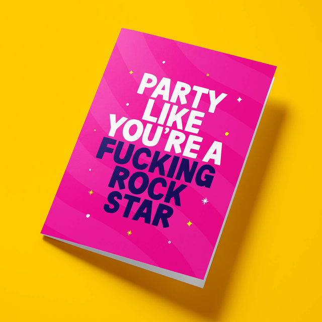 Hot pink 'Party Like A Rock Star' rude birthday card laid flat at an angle on a bright background, with dark navy 'FUCKING ROCK STAR' lettering standing out against the white text.