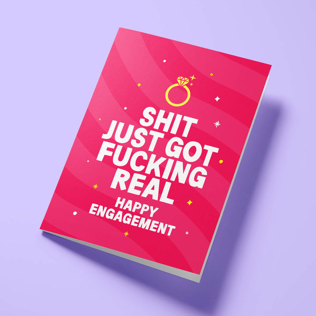 Raspberry pink 'Shit Just Got Real' rude engagement card laid flat at an angle on a bright background, with a gold diamond ring above bold white 'SHIT JUST GOT FUCKING REAL' lettering.