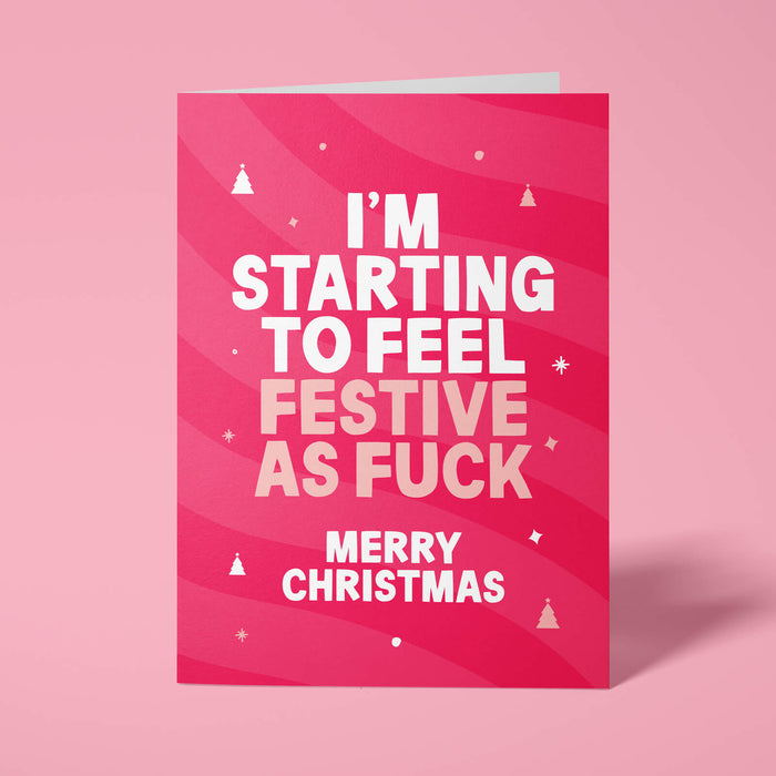 Raspberry pink rude Christmas card standing upright on a bright background. Bold white and pale pink chunky typography reads 'I'M STARTING TO FEEL FESTIVE AS FUCK MERRY CHRISTMAS' over pink wave stripes with white and pale pink Christmas tree and sparkle details.