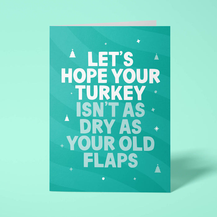 Green rude Christmas card standing upright on a bright background. Bold white and pale mint chunky typography reads 'LET'S HOPE YOUR TURKEY ISN'T AS DRY AS YOUR OLD FLAPS' over Green wave stripes with white Christmas tree and sparkle details.