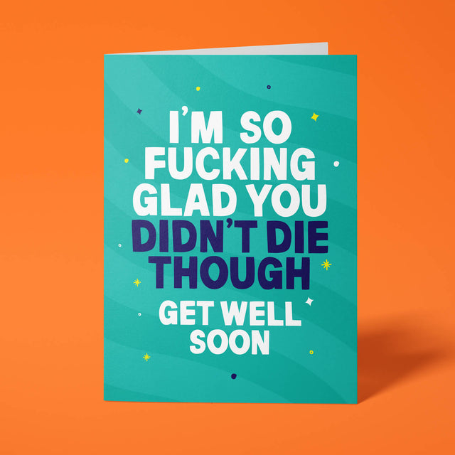 Teal rude get well soon card standing upright against a bright orange background. Bold white and dark navy chunky typography reads 'I'M SO FUCKING GLAD YOU DIDN'T DIE THOUGH GET WELL SOON' over soft teal wave stripes with yellow and navy sparkle and dot details.