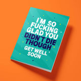 Teal 'Glad You Didn't Die' get well soon card laid flat at an angle on a bright orange background, with dark navy 'DIDN'T DIE THOUGH' lettering between white text.