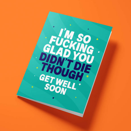 Teal 'Glad You Didn't Die' get well soon card laid flat at an angle on a bright orange background, with dark navy 'DIDN'T DIE THOUGH' lettering between white text.