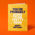 Golden yellow "You're Probably Going To Die Pretty Soon" birthday card tilted at an angle against a bold orange background