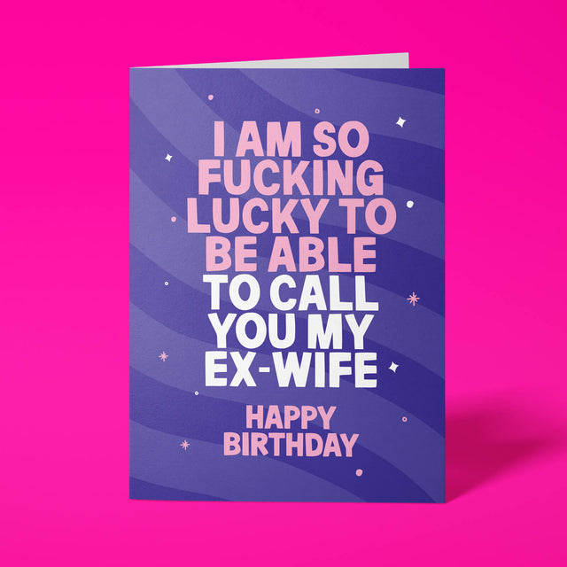 A birthday greeting card with a bold purple and blue striped background and pink and white text that reads, 'I AM SO FUCKING LUCKY TO BE ABLE TO CALL YOU MY EX-WIFE HAPPY BIRTHDAY,' shown standing against a bright pink background.