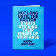 Sky-blue "Not Long Until The Doctor Starts Sticking Their Finger Up Your Arse" birthday card tilted at an angle against a deep blue background
