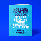 Sky-blue "Not Long Until The Doctor Starts Sticking Their Finger Up Your Arse" birthday card tilted at an angle against a deep blue background
