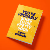A yellow greeting card with bold blue and white text reading 'YOU'RE PROBABLY GOING TO DIE PRETTY SOON' and 'HAPPY BIRTHDAY', shown at an angle on a bright orange background.