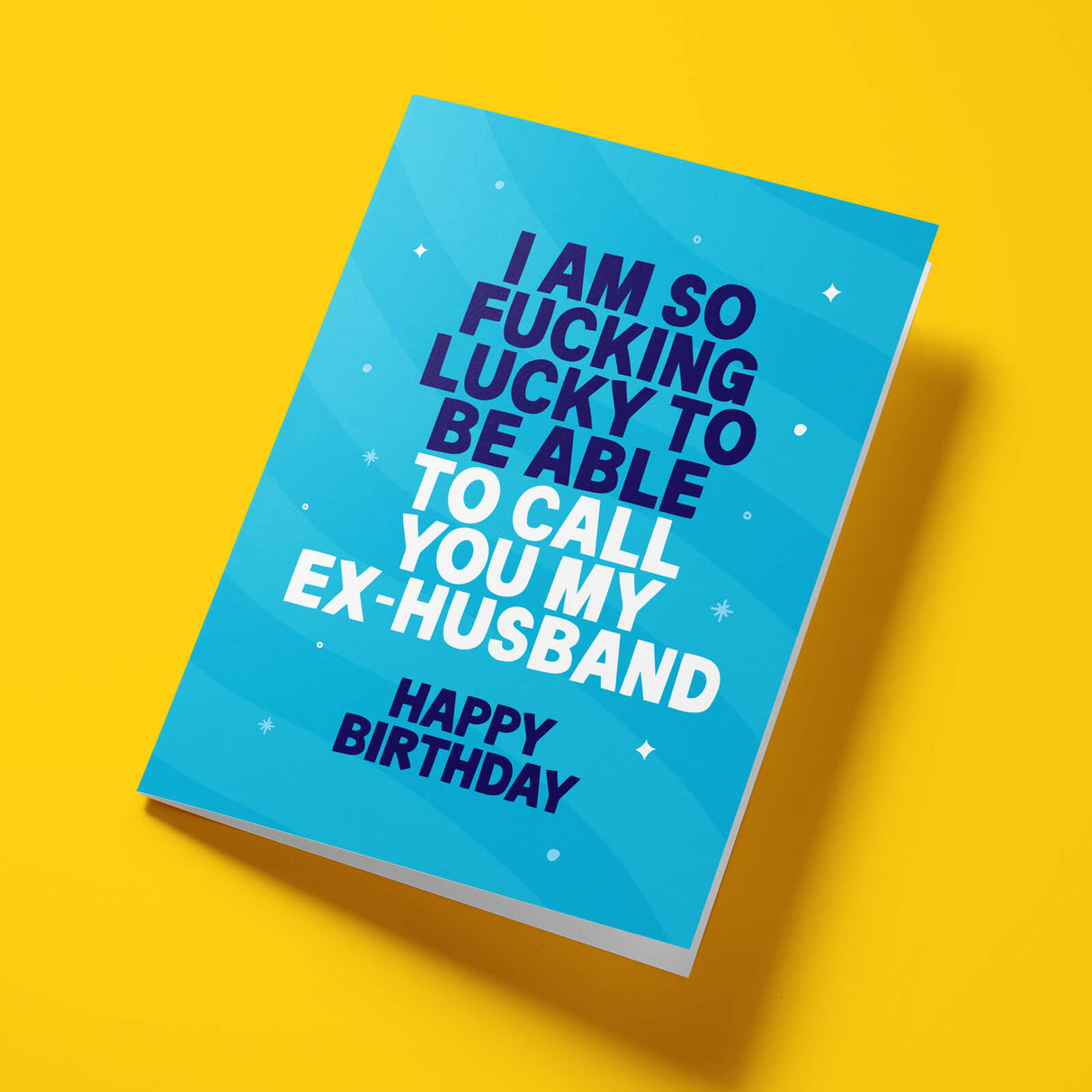A blue birthday card with bold text reading 'I AM SO FUCKING LUCKY TO BE ABLE TO CALL YOU MY EX-HUSBAND' and 'HAPPY BIRTHDAY' in white and dark blue, shown on a bright yellow background.