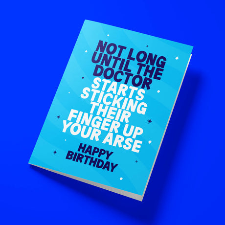 A blue greeting card with bold white and dark blue text reading 'NOT LONG UNTIL THE DOCTOR STARTS STICKING THEIR FINGER UP YOUR ARSE' and 'HAPPY BIRTHDAY', shown at an angle on a solid blue background.