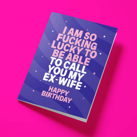 A greeting card with a bold purple and blue graphic design, featuring the text 'I AM SO FUCKING LUCKY TO BE ABLE TO CALL YOU MY EX-WIFE HAPPY BIRTHDAY' in pink and white lettering, displayed on a vibrant pink background.