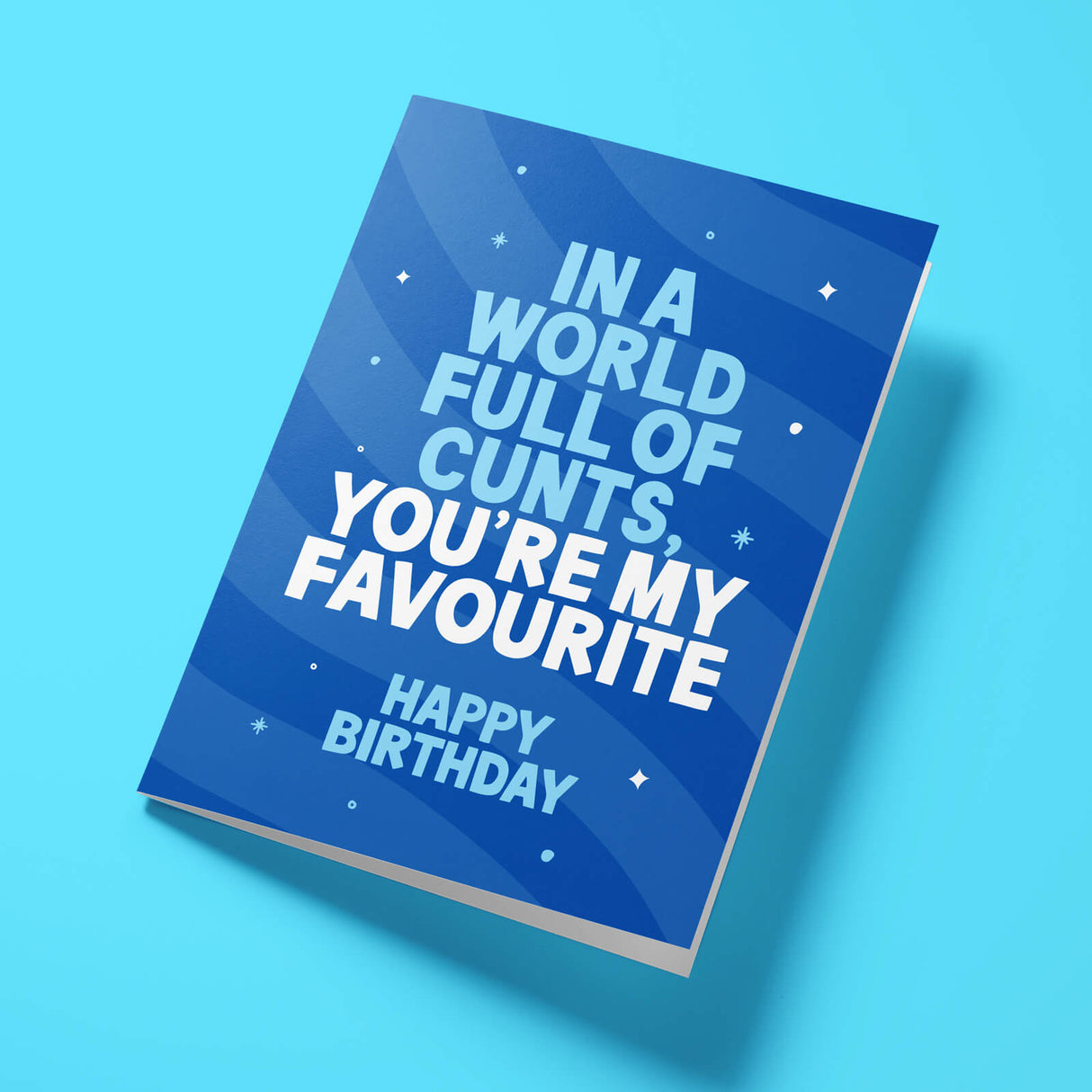 A blue birthday card with bold white text reading 'IN A WORLD FULL OF CUNTS, YOU'RE MY FAVOURITE HAPPY BIRTHDAY' and stars, shown standing at an angle on a light blue background.