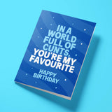 A blue birthday card with bold white text reading 'IN A WORLD FULL OF CUNTS, YOU'RE MY FAVOURITE HAPPY BIRTHDAY' and stars, shown standing at an angle on a light blue background.