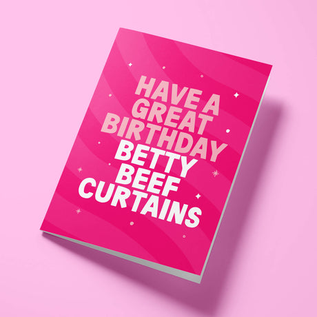 Hot pink Betty Beef Curtains rude birthday card for her laid flat at an angle on a pale pink background, showing bold white 'BETTY BEEF CURTAINS' lettering beneath pale pink 'HAVE A GREAT BIRTHDAY' text.