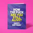 Deep purple A5 greeting card standing upright against a hot pink background. White chunky typography reads 'HOW THE FUCK' with bold bright yellow text below reading 'ARE YOU STILL ALIVE?', finished with white 'HAPPY BIRTHDAY'. Swirling wave pattern in darker purple tones with yellow dot and star details.