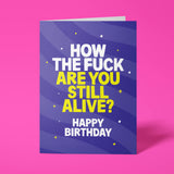 Deep purple A5 greeting card standing upright against a hot pink background. White chunky typography reads 'HOW THE FUCK' with bold bright yellow text below reading 'ARE YOU STILL ALIVE?', finished with white 'HAPPY BIRTHDAY'. Swirling wave pattern in darker purple tones with yellow dot and star details.