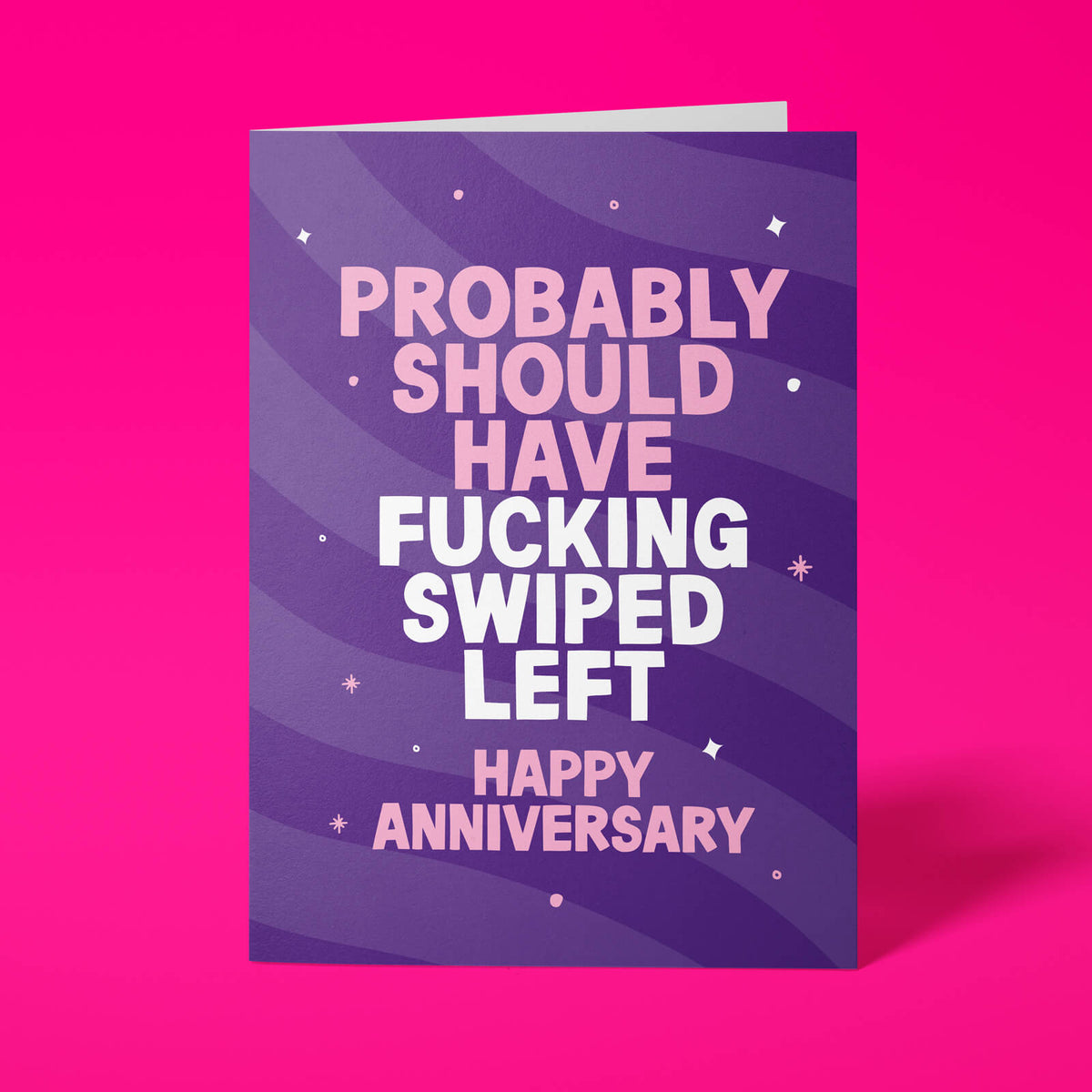 Deep purple anniversary card with pink and white block-capital text reading "PROBABLY SHOULD HAVE FUCKING SWIPED LEFT, HAPPY ANNIVERSARY," decorated with hand-drawn stars, dots and sparkle icons.