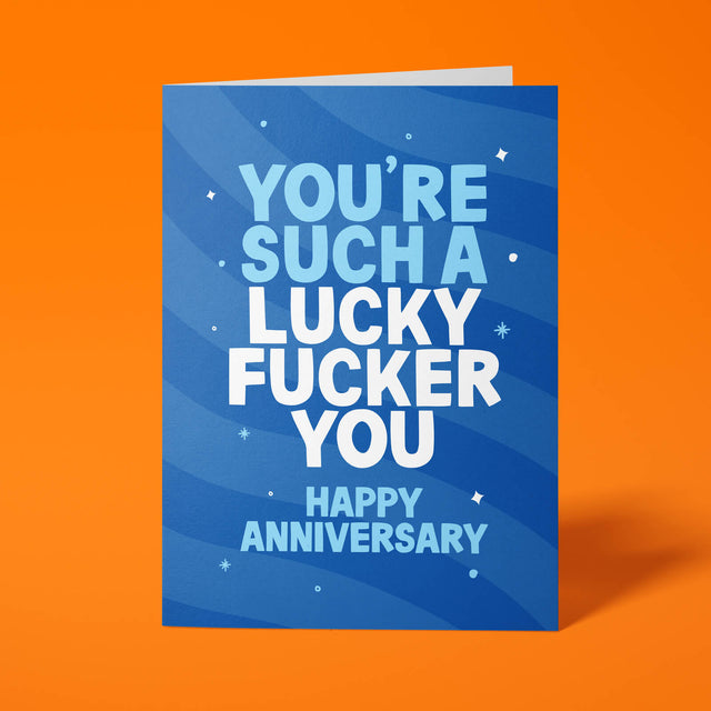 Royal blue anniversary card with pale blue and white block-capital text reading "YOU'RE SUCH A LUCKY FUCKER YOU, HAPPY ANNIVERSARY," decorated with hand-drawn stars, dots and sparkle icons.